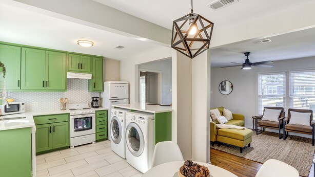 A modern kitchen with green cabinets, a white countertop, and a built-in washing machine. There is a dining area with a table and chairs, and a living room with a yellow sofa and brown chairs. - 2 Blocks to At&t Stadium: Sports Fan Headquarters! (Arlington)