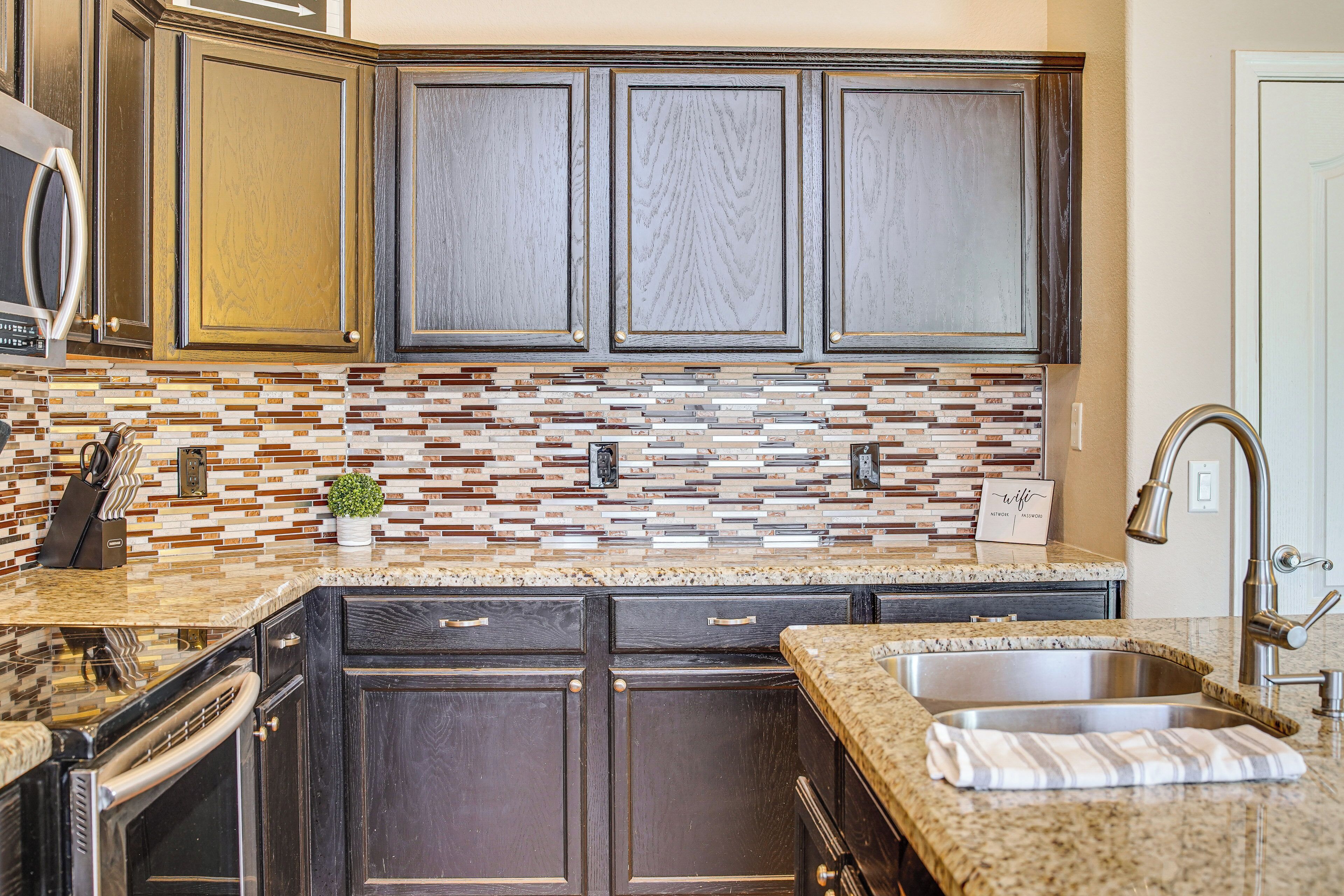 Una cocina con gabinetes de madera oscura, salpicadero de azulejos, encimeras de granito, un lavamanos de acero inoxidable y un horno empotrado.