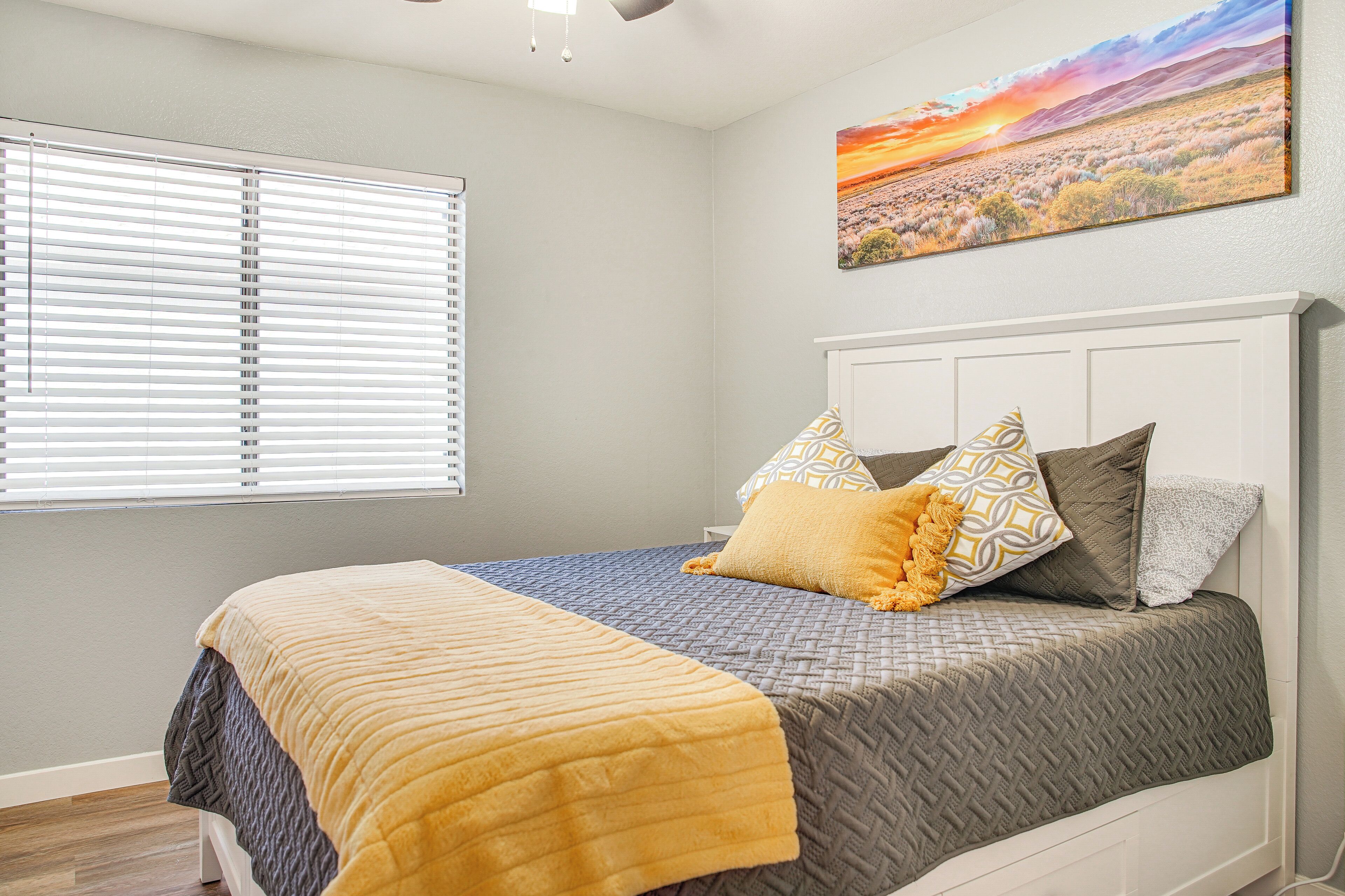 A bedroom with a bed, pillows, a blanket, a painting, and a window with blinds.