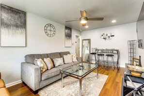 A modern living room with a gray sofa, a glass coffee table, wooden flooring, a ceiling fan, and a wall clock.