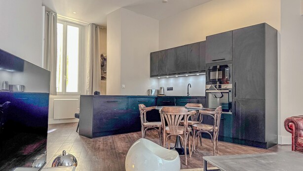 A modern kitchen with dark cabinets, a central island, and a dining area with wooden chairs and a white bowl.