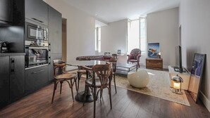 A modern kitchen with dark cabinetry, a dining area with wooden chairs and a round table, and a living space with a sofa and a painting.