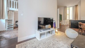 A modern living room with a flat-screen TV on a white stand, a dining area with a wooden table and chairs, and a staircase leading to an upper level.