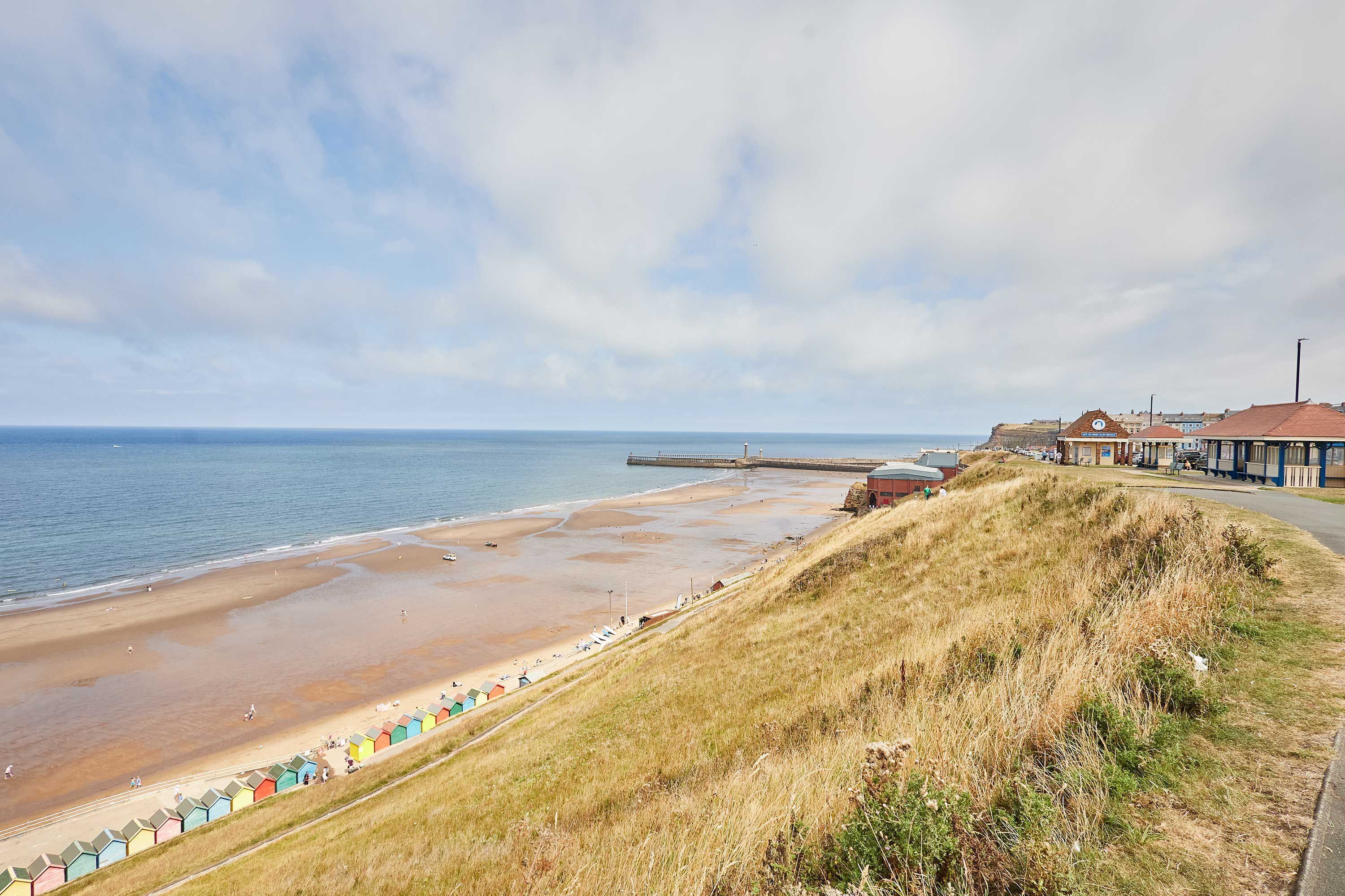 A coastal beach with a grassy embankment, a pier, and colorful beach huts along the shore.