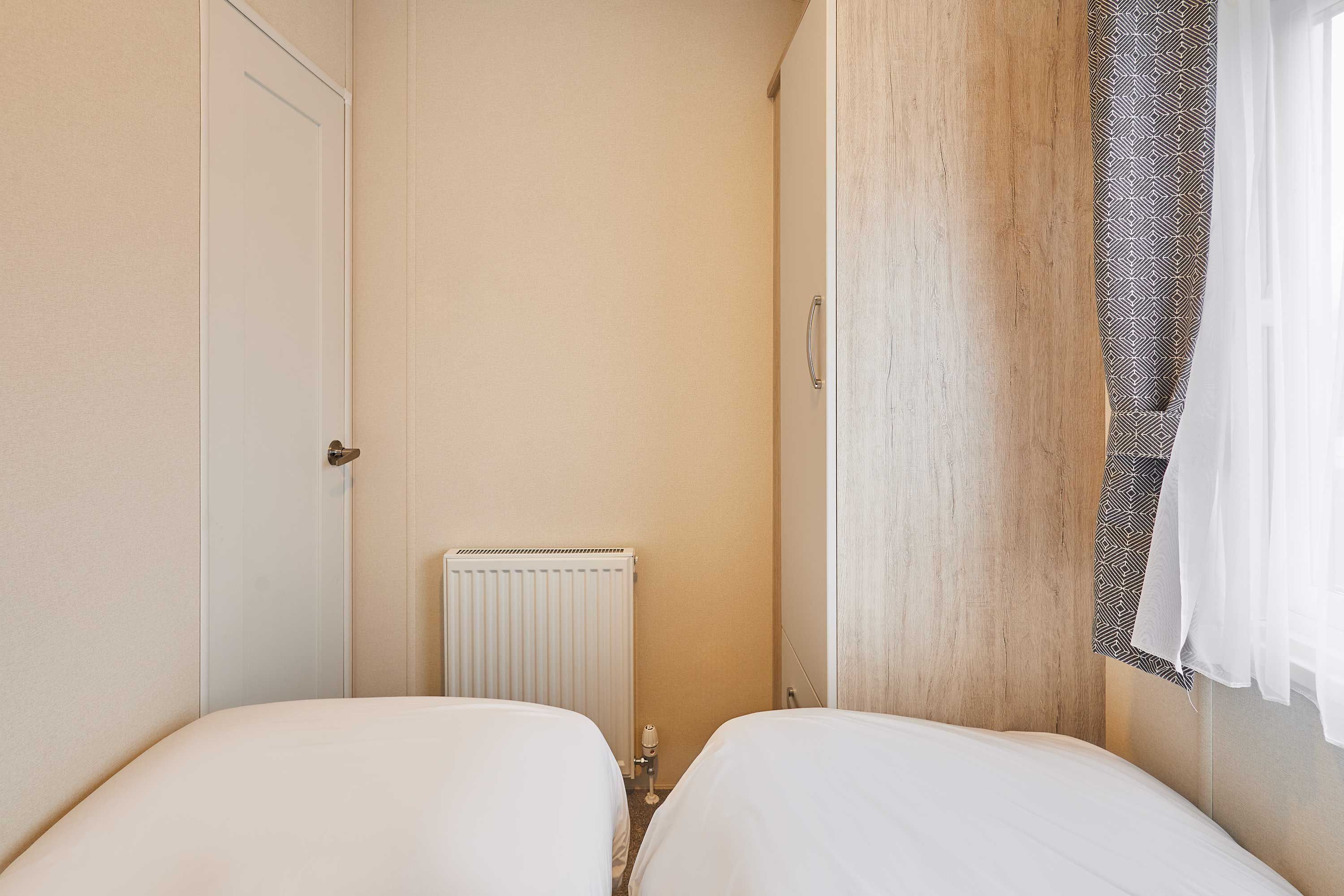 A small, narrow room with two beds, a radiator, and a window with patterned curtains.