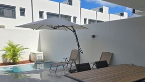 Et poolområde med et træbord og to liggestole, en stor parasol og en varmtvandsbeholder.