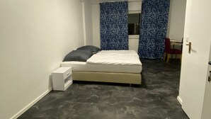 Ein kleiner, schmaler Raum mit einem Bett, einem Schreibtisch und einem Stuhl. Der Boden ist dunkel, die Wände weiß.