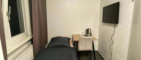 A small, single-bed room with a desk and chair, a window with curtains, a radiator, and a mounted TV.