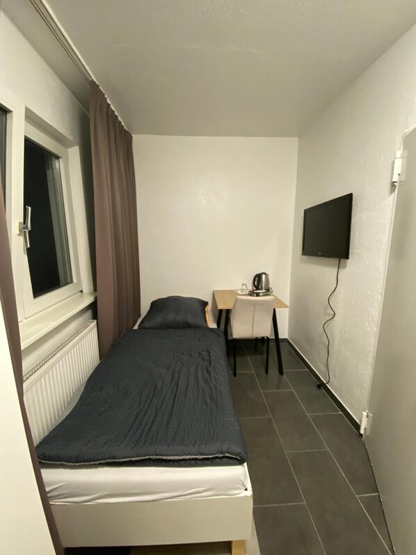 Una camera piccola con un letto singolo, una scrivania e una sedia, una finestra con tende, un termosifone e una TV a parete.