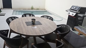 A round wooden table with black chairs, a grill, and a pool in the background. - Soleil & Sage Haven,2BR Townhouse in Cupecoy (Cupercoy)