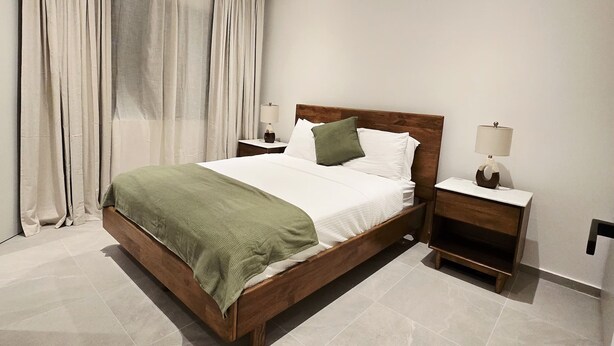 A modern bedroom with a wooden bed, white linens, a green throw, and a nightstand with a lamp.