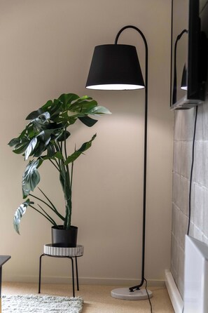 A potted plant on a small stool next to a floor lamp in a room with a tiled wall and a carpeted floor.