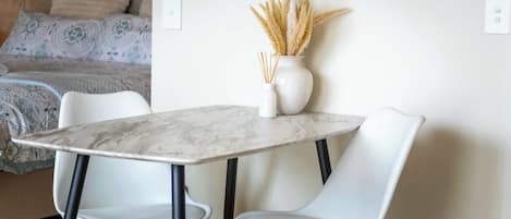 Una habitación moderna con una mesa de mármol, dos sillas blancas, un florero con plantas secas y una cama con ropa de cama estampada.