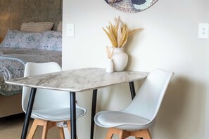 Une chambre moderne avec une table en marbre, deux chaises blanches, un vase contenant des plantes séchées et un lit avec une literie à motifs.