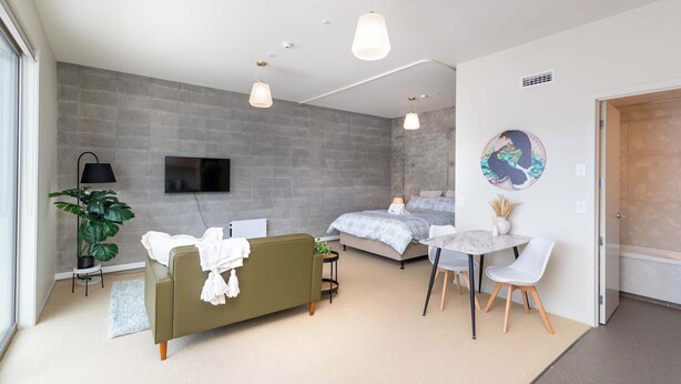 A modern bedroom with a bed, a sofa, a desk, and a TV. - Petone Studio | Balcony | Near Beach + Shops (Lower Hutt)