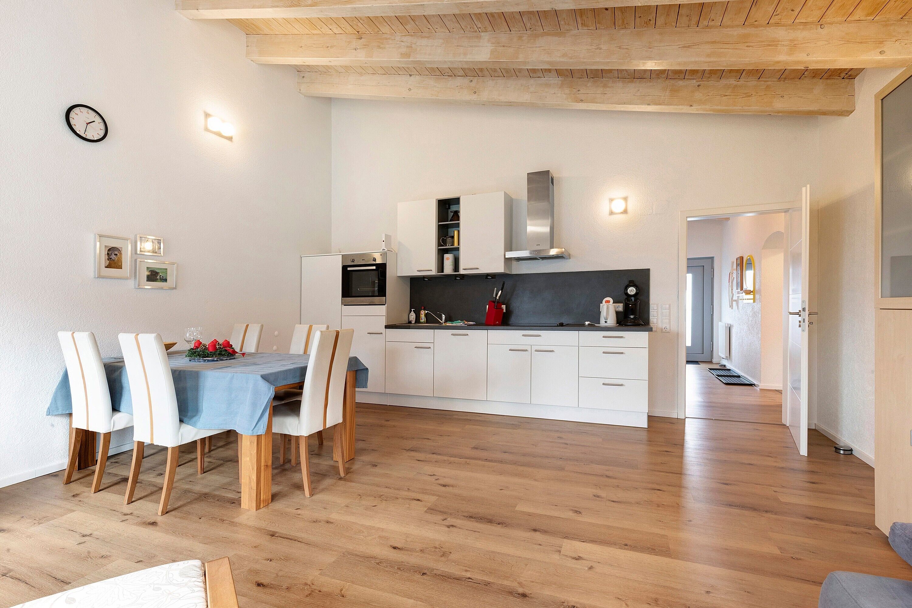 Een moderne keuken met witte kasten, een zwart aanrechtblad en een roestvrijstalen afzuigkap. Er is een eethoek met een tafel en stoelen, en een houten vloer door de hele ruimte.