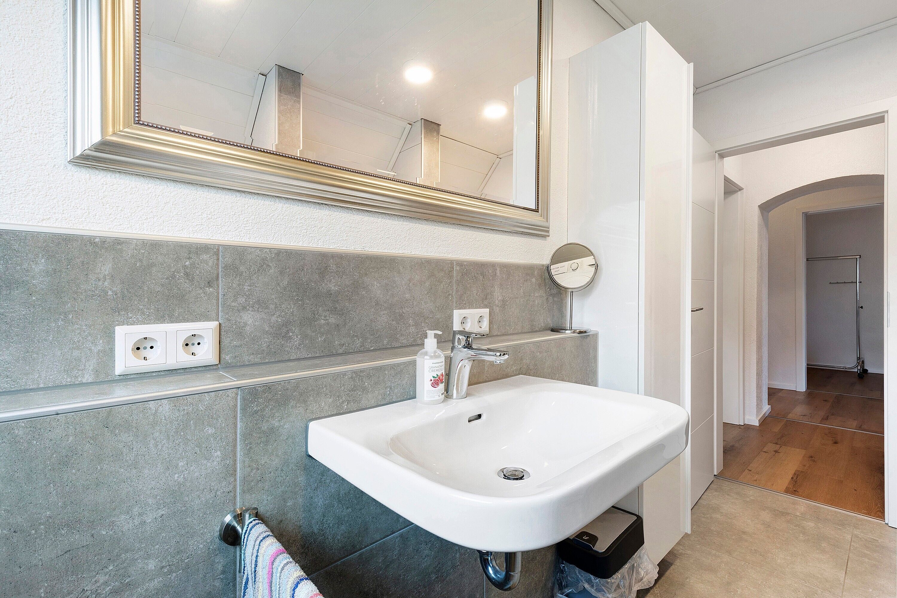 Un baño con un lavamanos blanco, un espejo y un perchero para toallas.