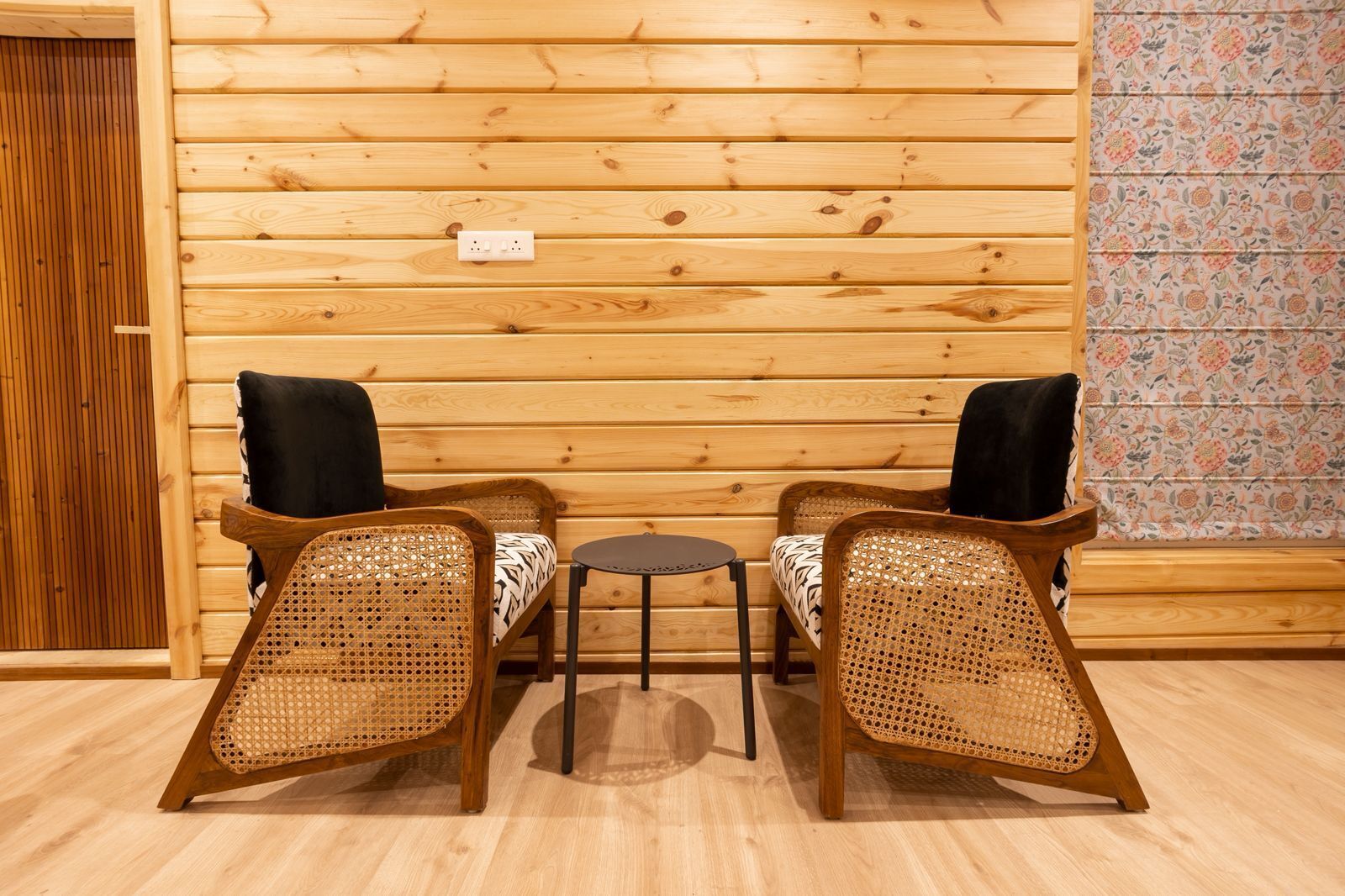 Interior de una cabaña de madera con dos sillones de mimbre, una mesita redonda y una pared de madera con toma de corriente.