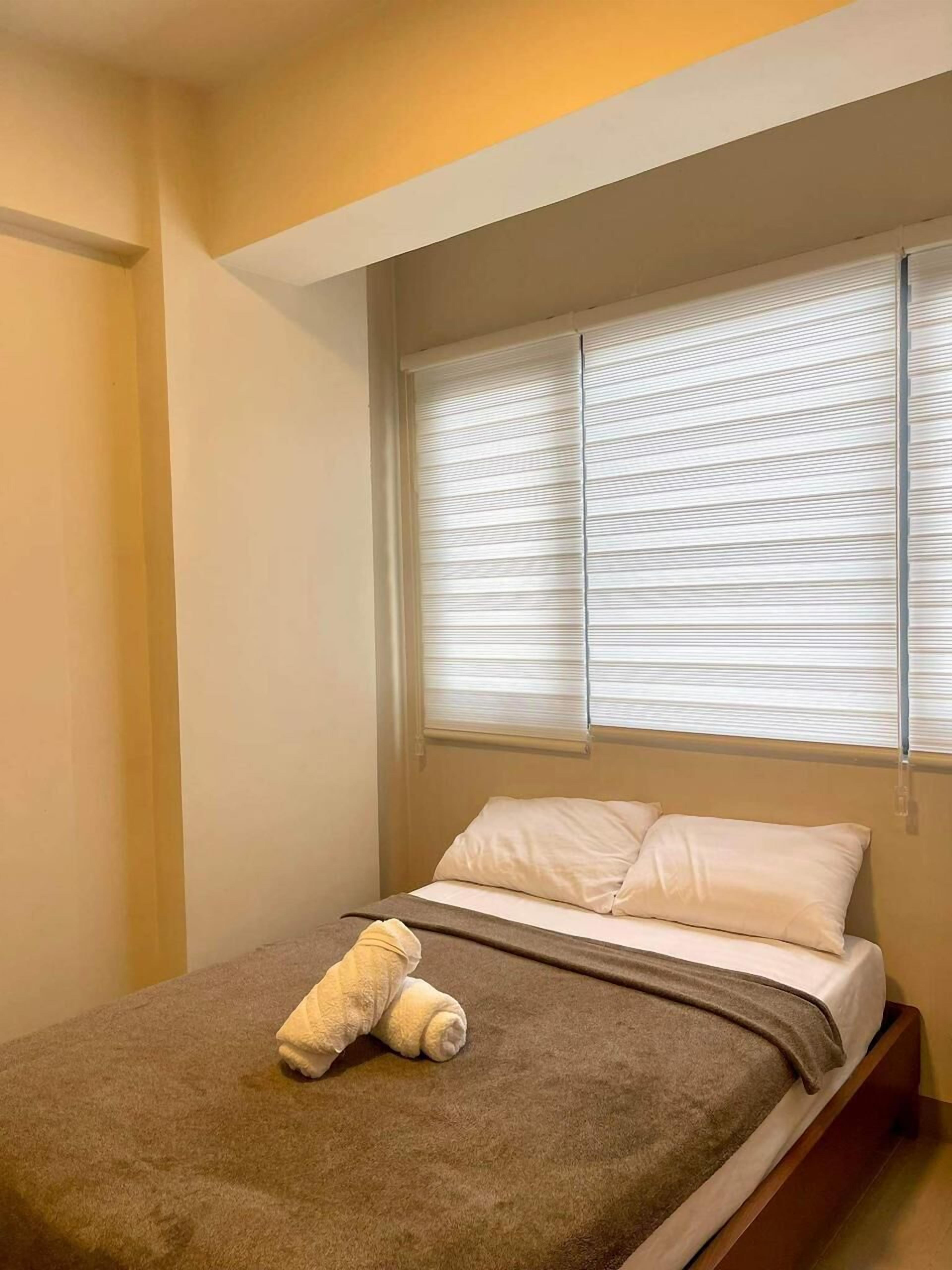 Una cama individual con un edredón marrón, almohadas blancas y un osito de peluche blanco sobre la cama.