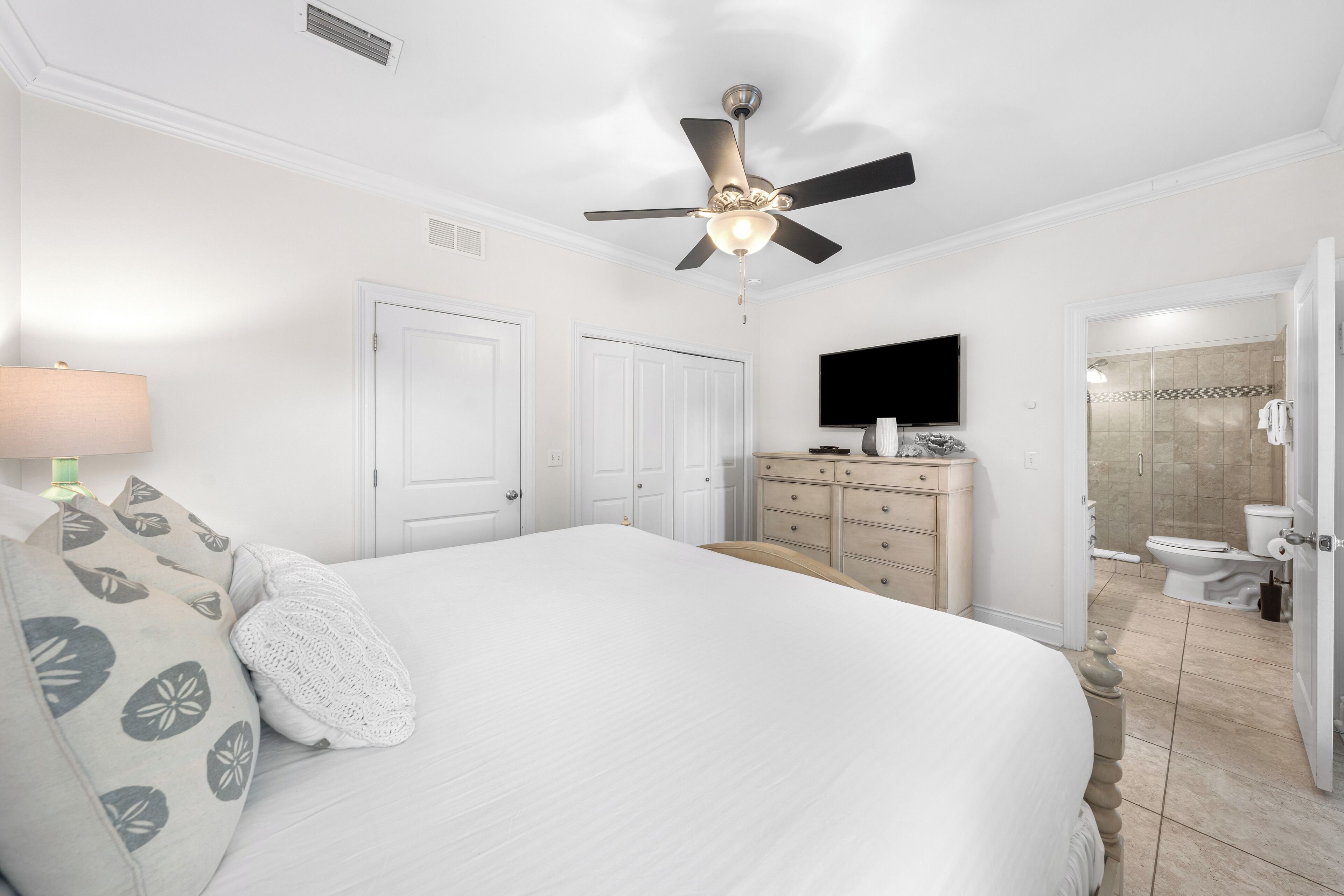 A bedroom with a bed, a ceiling fan, a dresser, a television, and a bathroom with a shower and toilet.