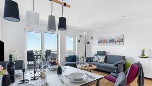 Ett modernt vardagsrum med en matplats. Här finns en soffa och ett matbord dukat med tallrikar, glas och bestick. En tavla hänger på väggen, och det finns stora fönster.