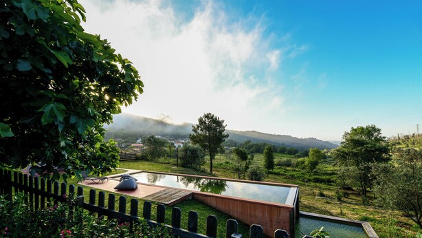 Área de piscina moderna al aire libre con muelle de madera y sillones de descanso, rodeada de vegetación exuberante y vistas a colinas distantes.