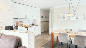 Een moderne keuken met witte kasten, een eethoek met een houten tafel en grijze stoelen, en een zithoek met een leren bank.
