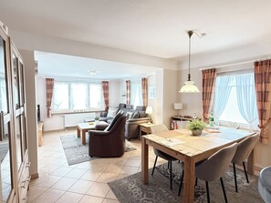 A spacious living room with a sofa, a dining table with chairs, and a kitchen area in the background. - Comfortable vacation apartment in Haus Frida-Sophie with a wood-burning stove, terrace, and garden (Sylt)