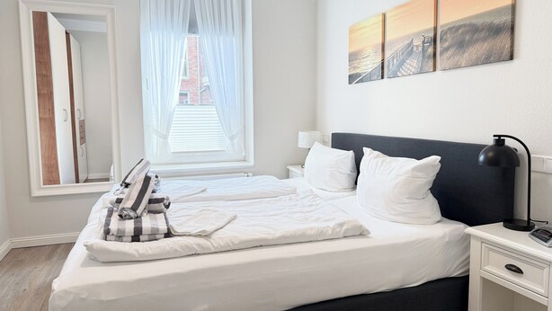 Een net opgemaakt bed met witte lakens en een geruit plaid, een bedlampje en een nachtkastje met een lamp.