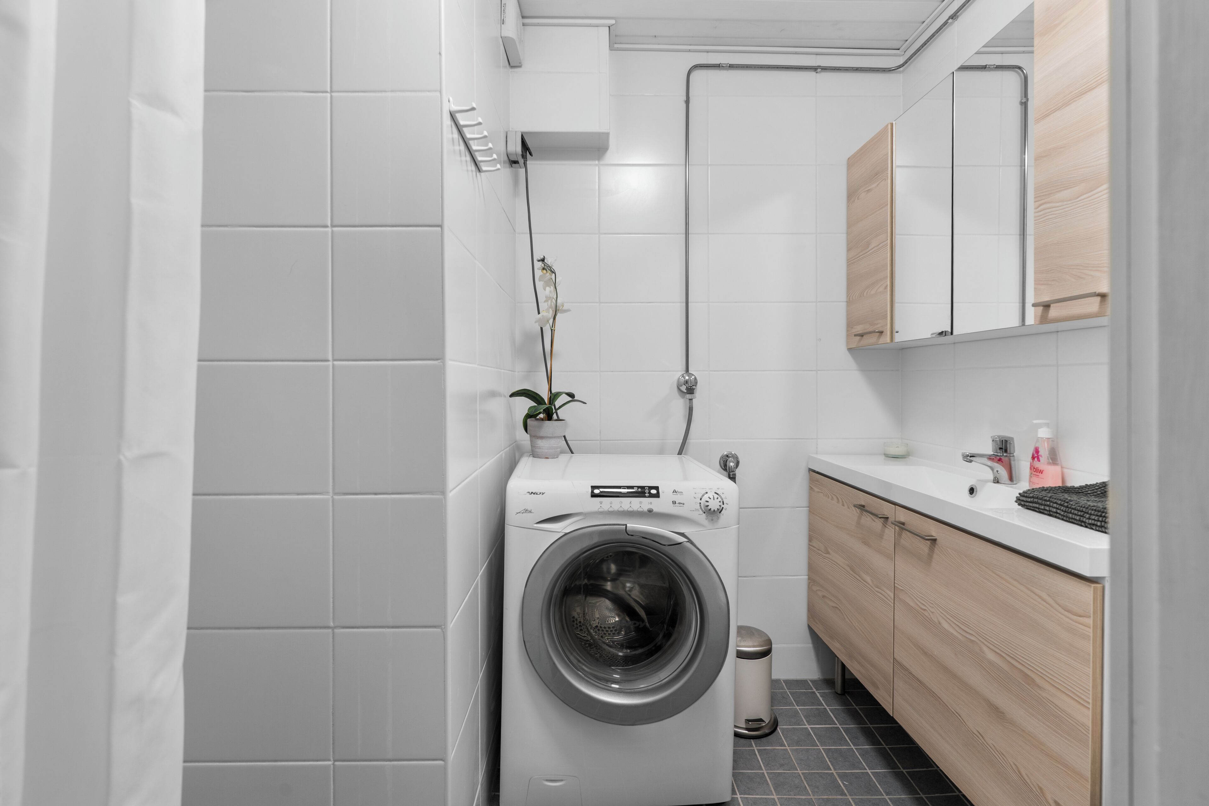 Un baño moderno y compacto con lavadora de carga frontal, un pequeño cubo de basura y un mueble de tocador de madera con espejo.