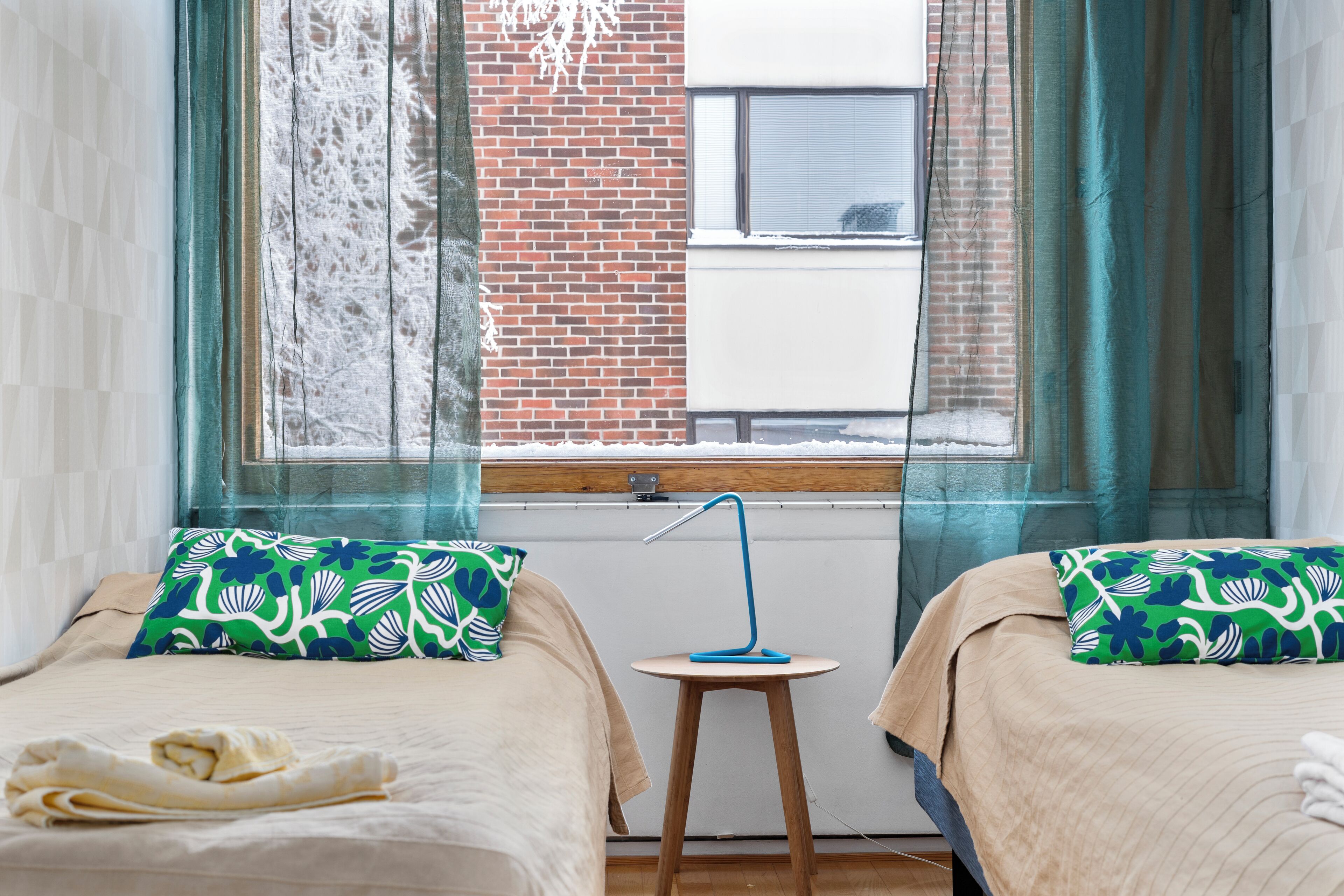 Ein Hotelzimmer mit zwei Betten, einem Fenster mit Vorhängen, einem Heizkörper und einem kleinen Tisch mit Lampe.