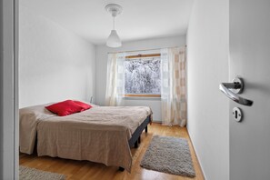 Um quarto com cama, janela com cortinas, porta com maçaneta e piso de madeira.