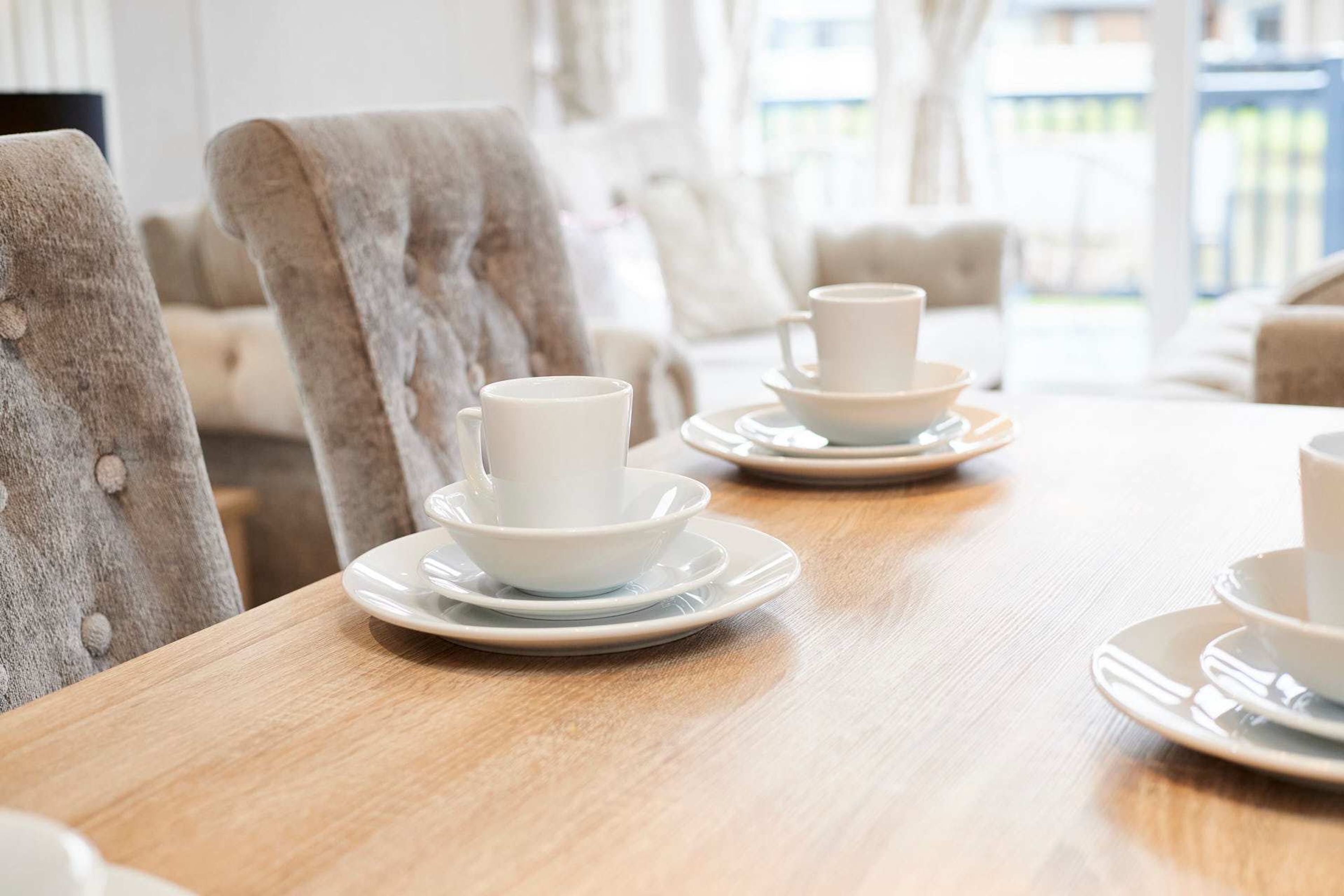A dining table set with white cups and saucers, upholstered chairs, and a wooden table surface.