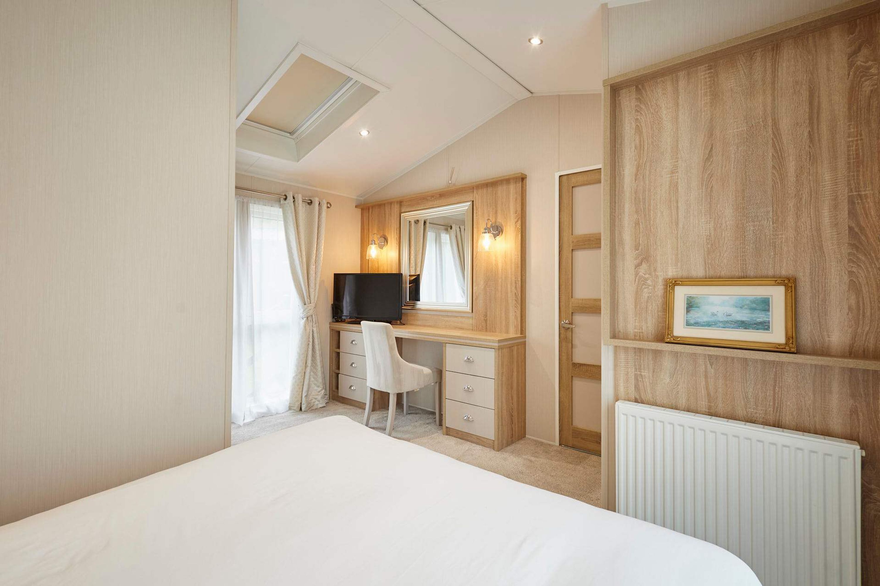 A hotel bedroom with a bed, a desk with a chair, a television, a mirror, and a framed picture on the wall.