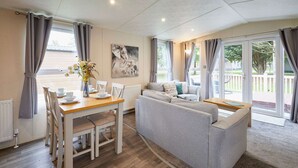 Ein modernes Wohnzimmer mit Essbereich, einem grauen Sofa, einem Holzesstisch und großen Fenstern mit Vorhängen.