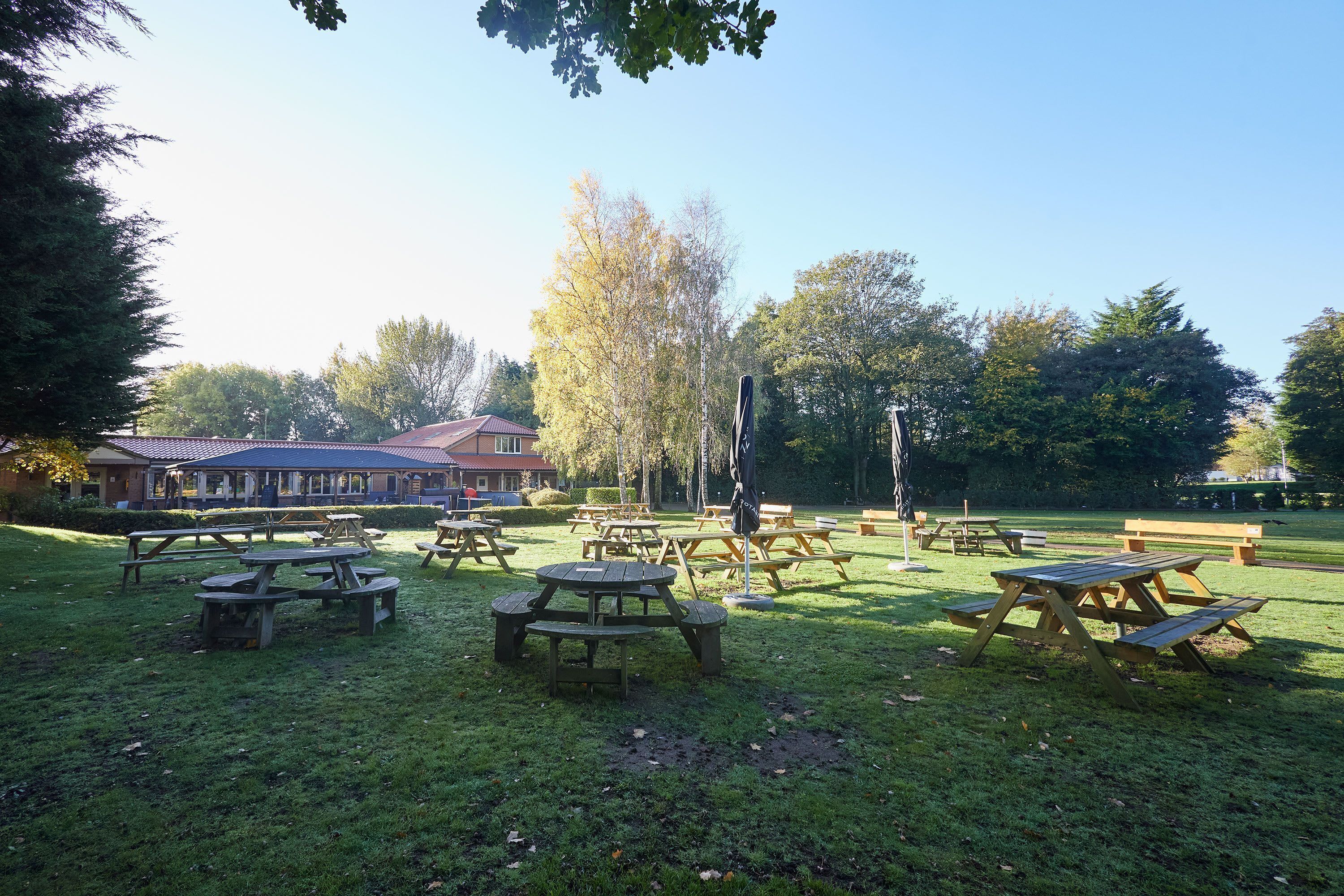 A park with picnic tables and benches, surrounded by trees and a building in the background.