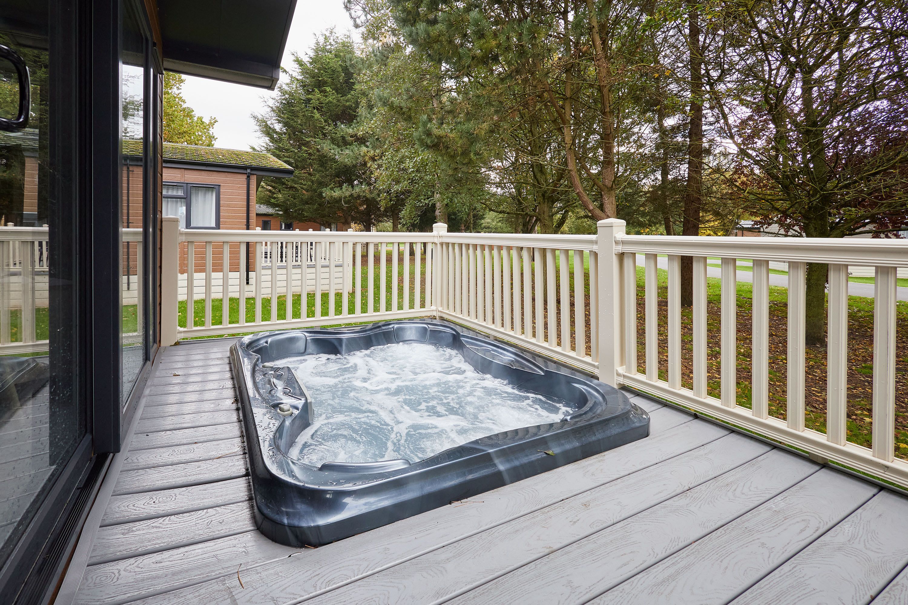 A hot tub on a wooden deck with a white fence and trees in the background.