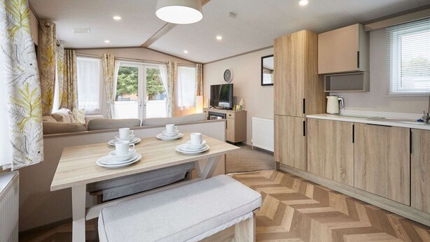 Une cuisine moderne avec un parquet en bois disposé en chevrons, une table à manger avec un banc, et un téléviseur posé sur un meuble.