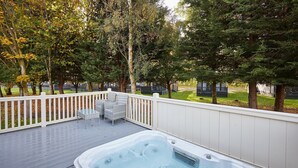 Un bain à remous sur une terrasse en bois, avec une clôture blanche et des chaises, le tout entouré d’arbres.
