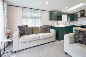 A modern living room with a sofa and armchair, a small table with a lamp, and a kitchen area with green cabinets and a window. - Host Stay Teejay s Lodge (York)