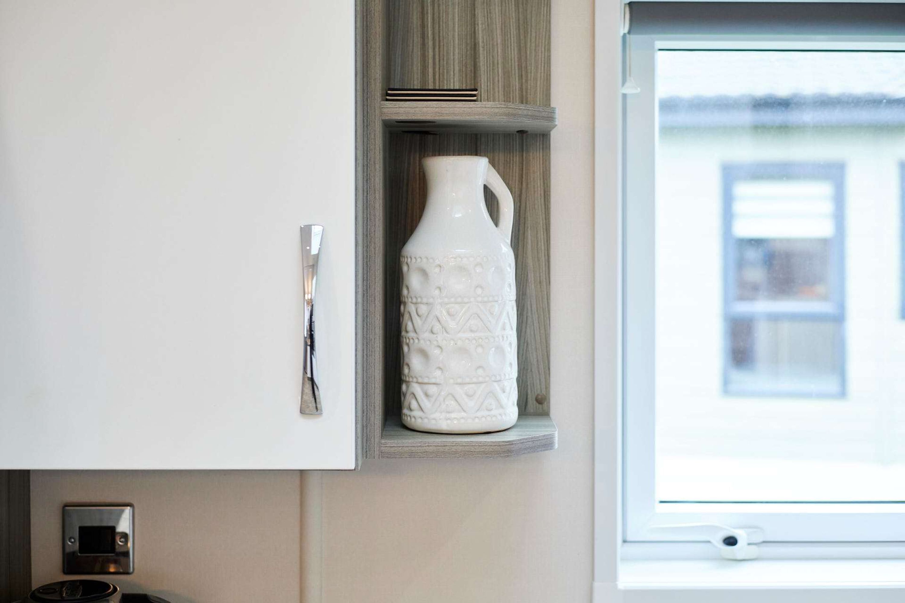 A white cabinet with a handle, a white ceramic pitcher with a handle, and a window with a view of a building outside.