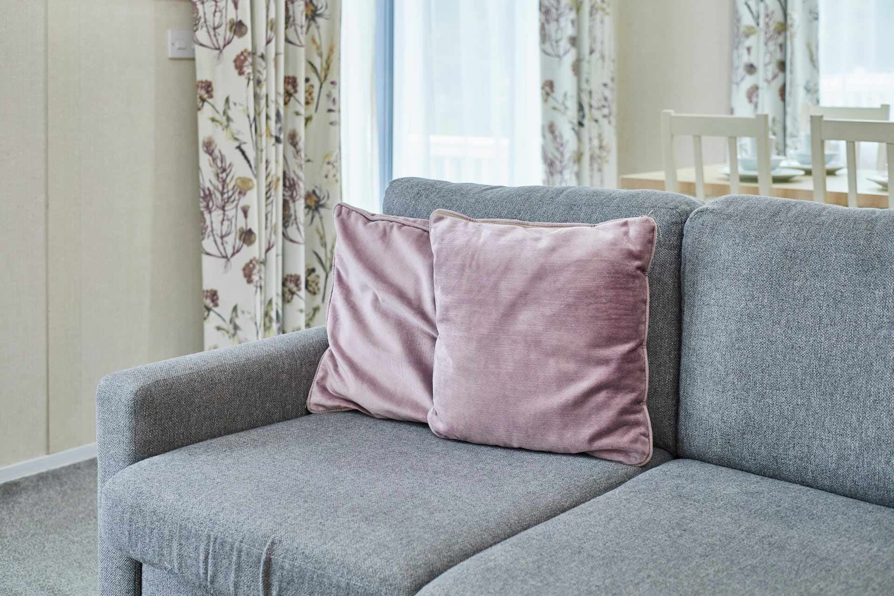 Un sofá gris con dos cojines rosados en una habitación con cortinas florales y una silla blanca al fondo.