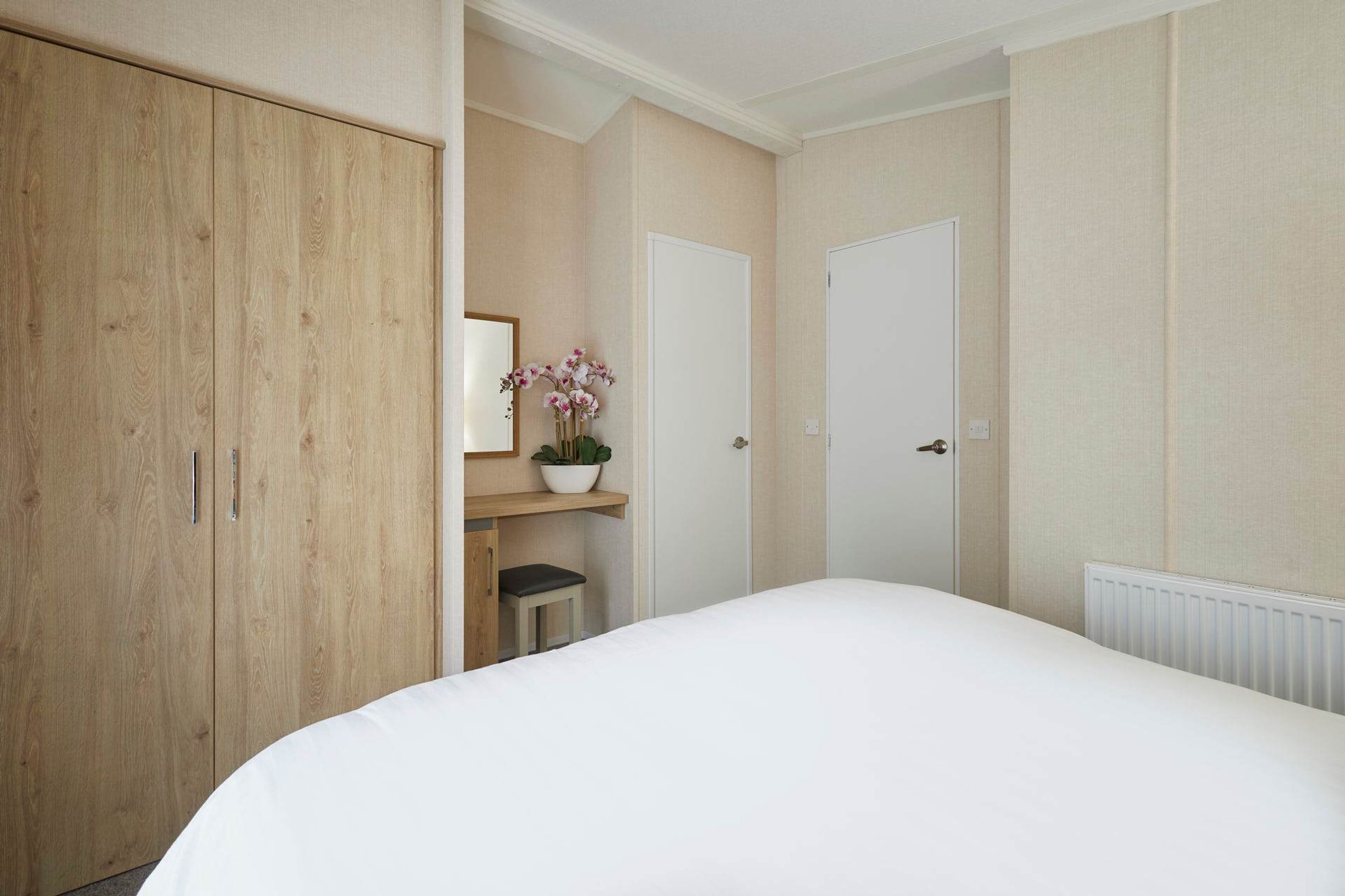 A bedroom with a large bed, wooden wardrobe, a small table with a potted plant, and two white doors.