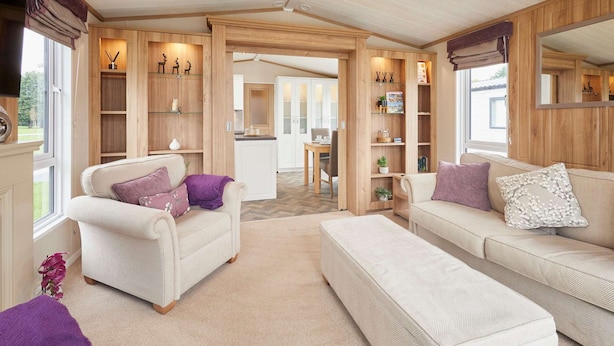Een gezellige woonkamer met een bank, een fauteuil en een salontafel. Er staan houten planken met decoratieve objecten en in de achtergrond een eethoek.