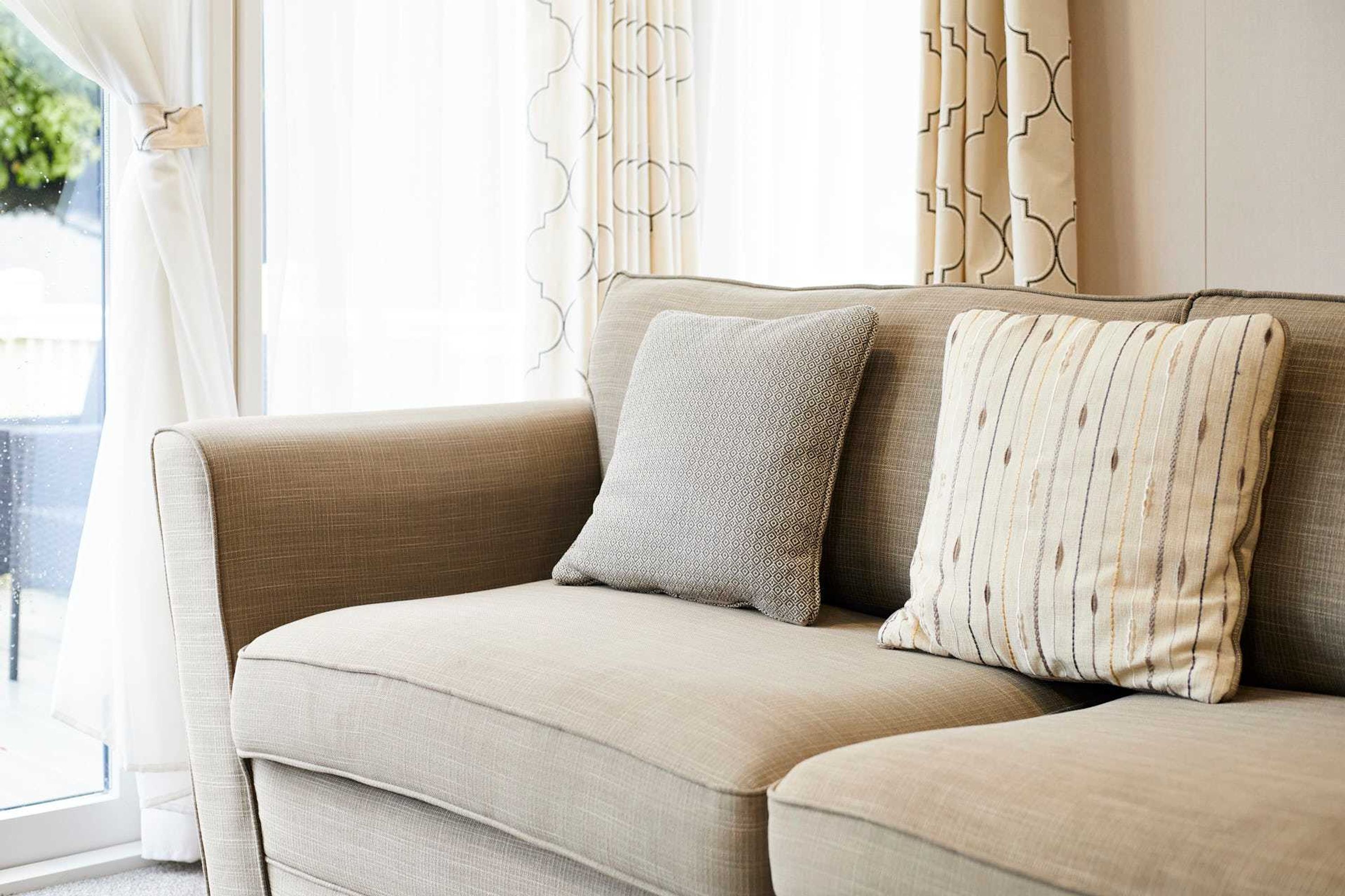 A beige sofa with two pillows, a window with curtains, and a view of greenery outside.