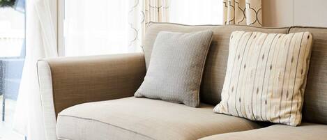 A beige sofa with two pillows, a window with curtains, and a view of greenery outside.