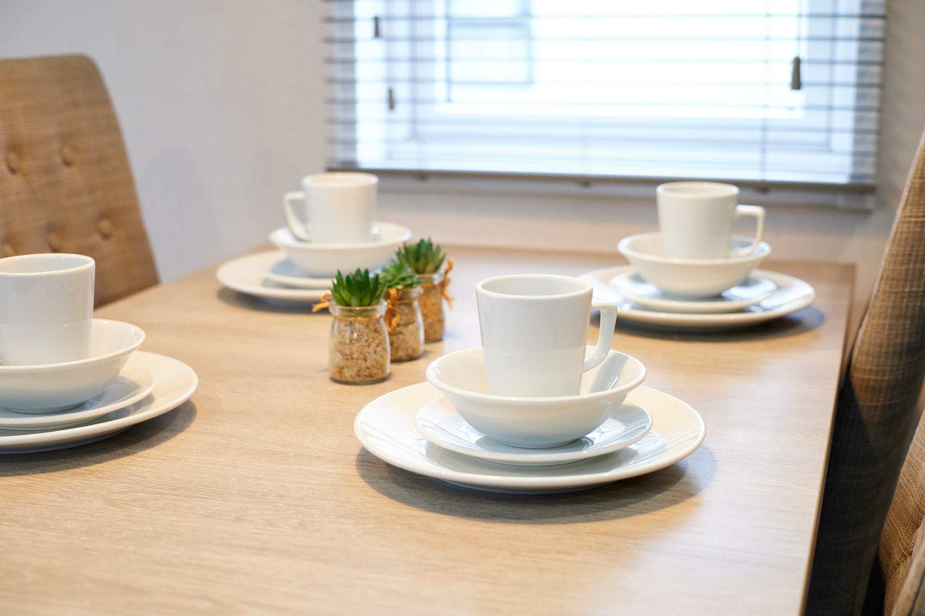A dining table with white cups and saucers, small potted plants, and a window in the background.