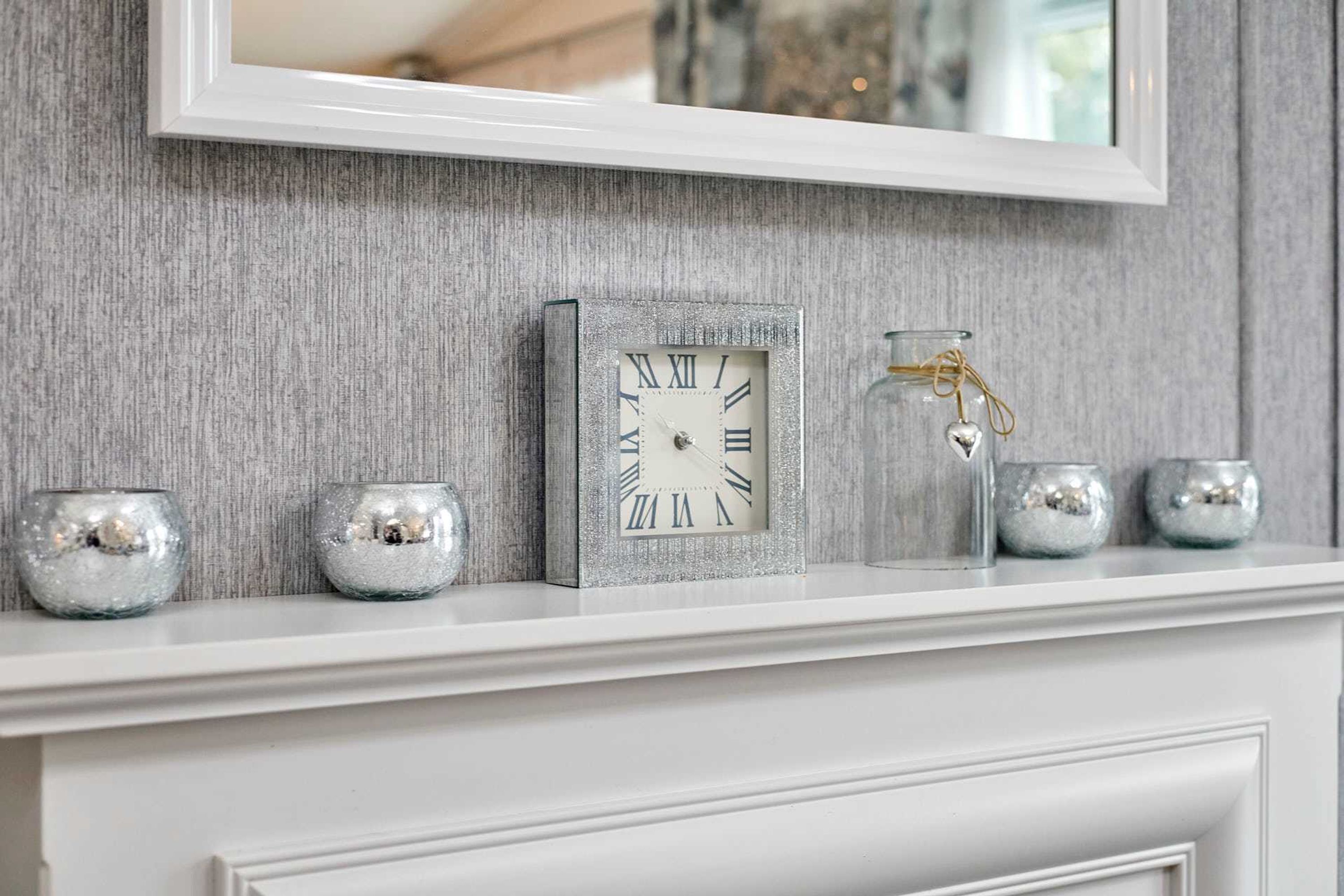 Une cheminée blanche ornée d’une horloge encadrée d’argent, de deux vases en verre décoratifs et de trois chandeliers en argent.