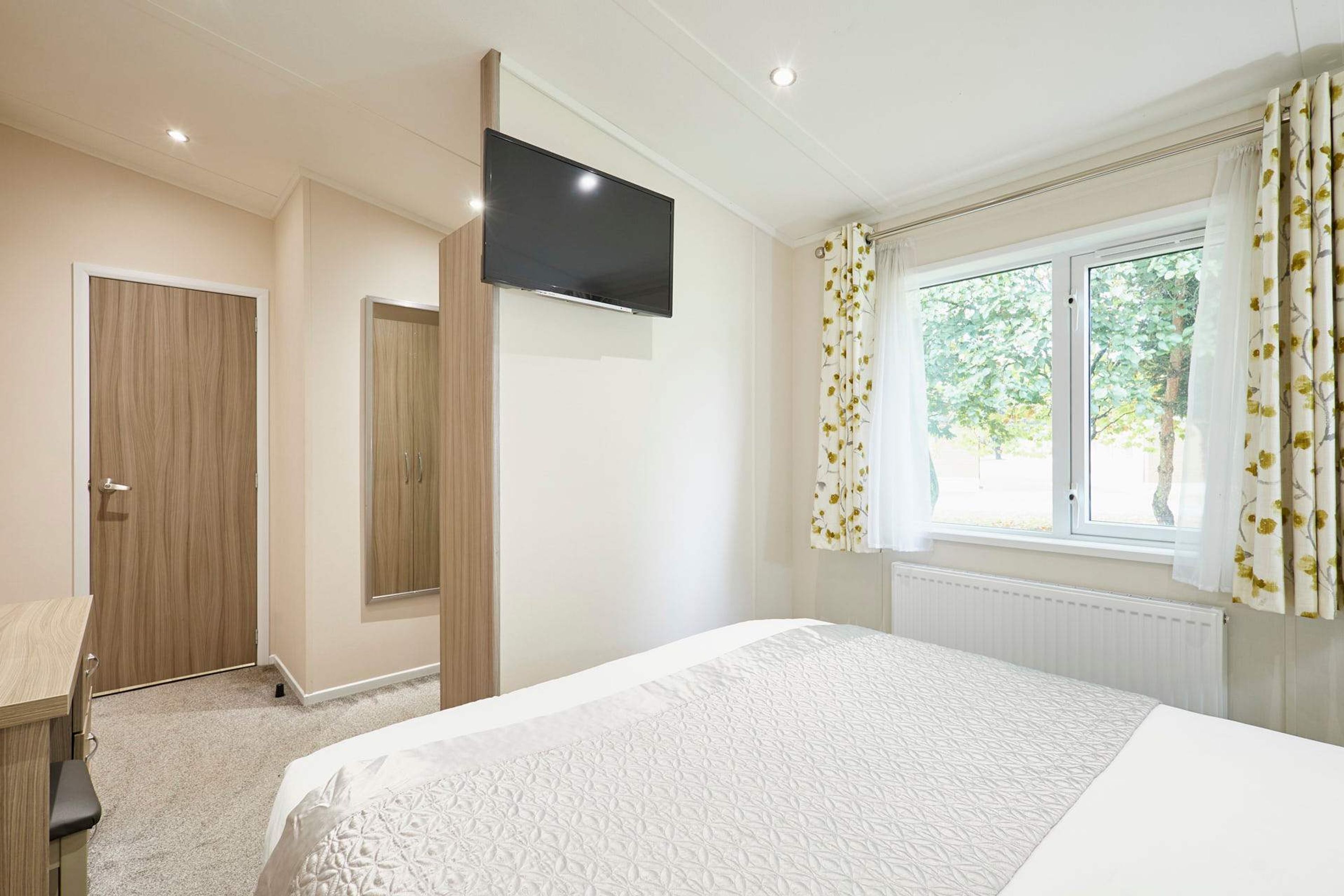 A bedroom with a bed, a TV mounted on the wall, a window with curtains, and a door.