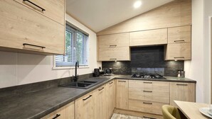 A modern kitchen with wooden cabinets, a black countertop, and a window with blinds. - Host Stay Fir Tree Lodge (York)